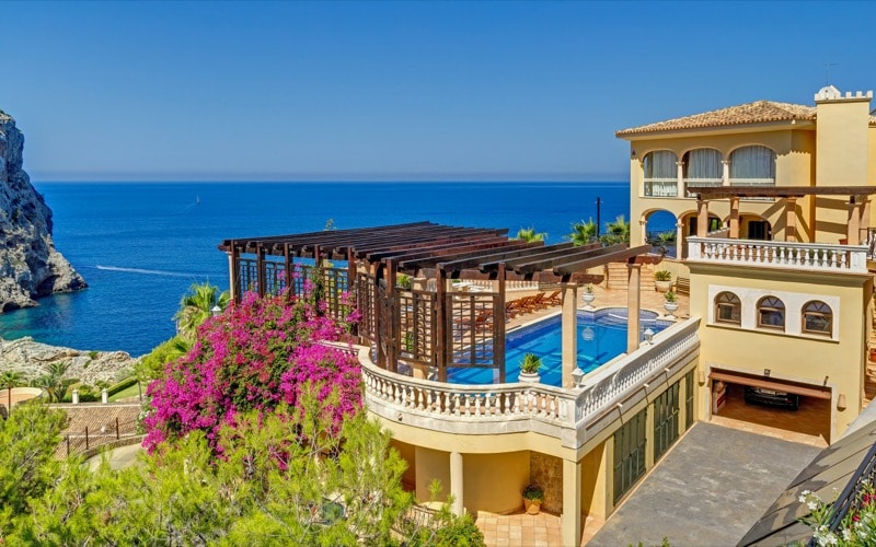 An image of a traditional sea view villa in Port Andratx, showcasing classic Mediterranean architecture with terracotta roofs, stone facades, and elegant arches. The villa is surrounded by lush gardens and offers breathtaking views of the Mediterranean Sea. Inside, the home combines rustic charm with modern comforts, featuring spacious rooms with natural light, cozy living areas, and stylish décor. The outdoor space includes a private pool, large terrace, and shaded seating areas, ideal for enjoying the stunning coastal landscape.