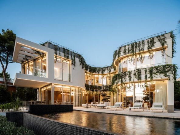 An image of a stunning architectural masterpiece located near the beach. The structure features bold, contemporary design with clean lines, expansive glass panels, and unique geometric elements. Surrounded by a meticulously landscaped garden, the home seamlessly blends into its coastal environment. In the background, the golden sands and azure waves of the nearby beach enhance the property's allure, reflecting its exclusive and sophisticated character.